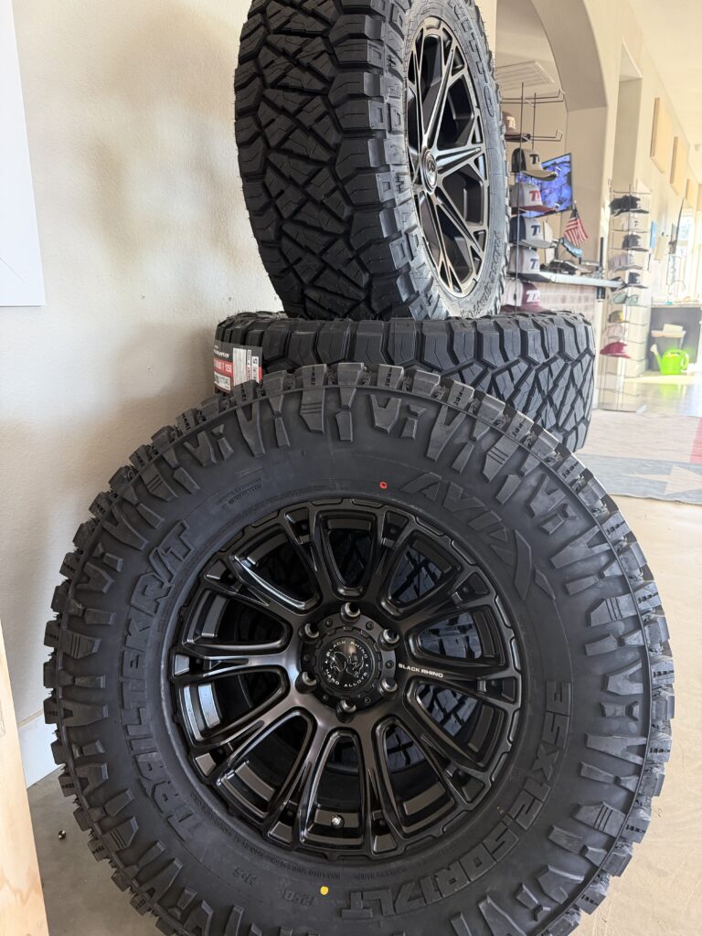 Black rhino wheel and tire displayed in front of a stack of tires.
